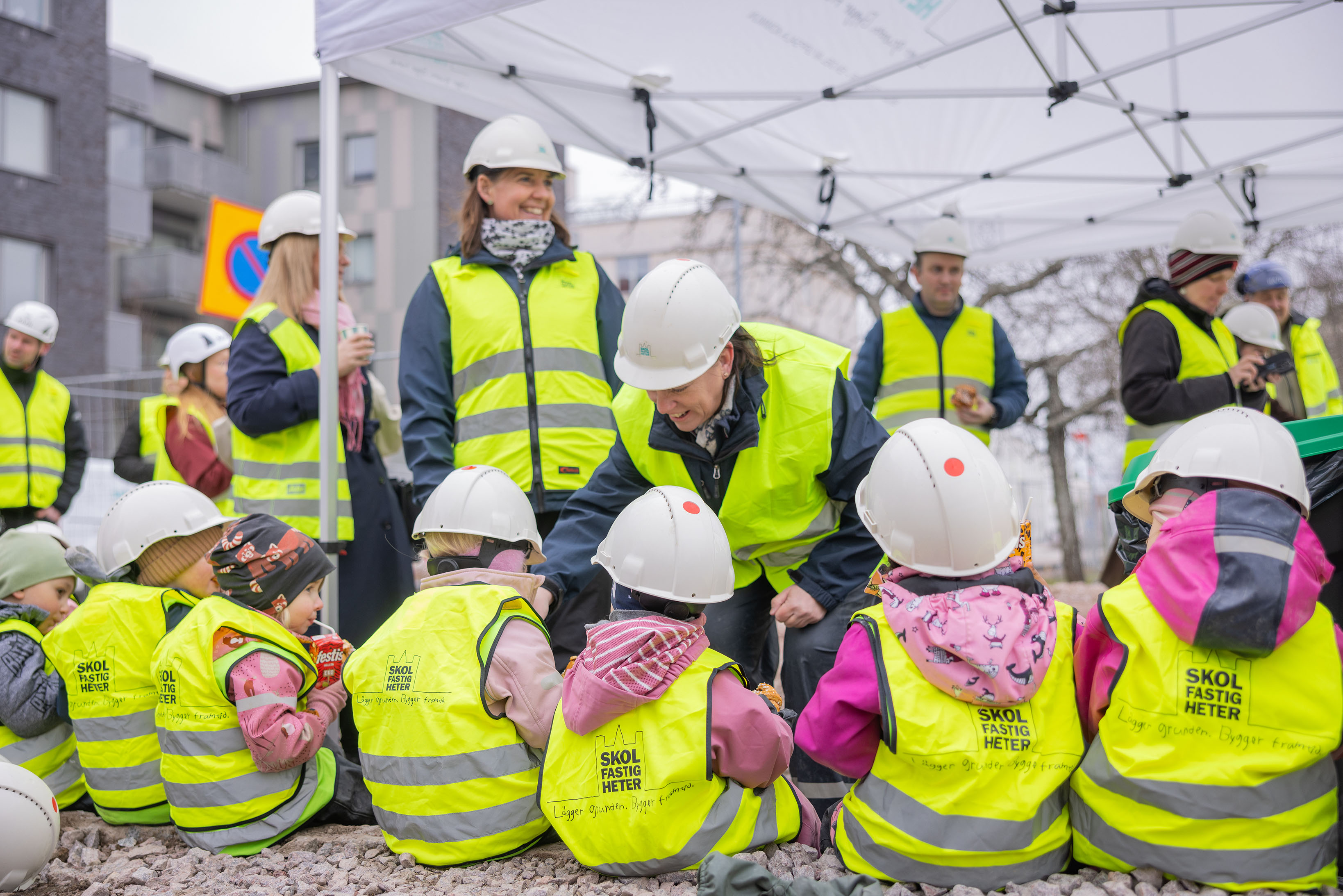 Förskolebarn i gula byggvästar sitter och fikar.
