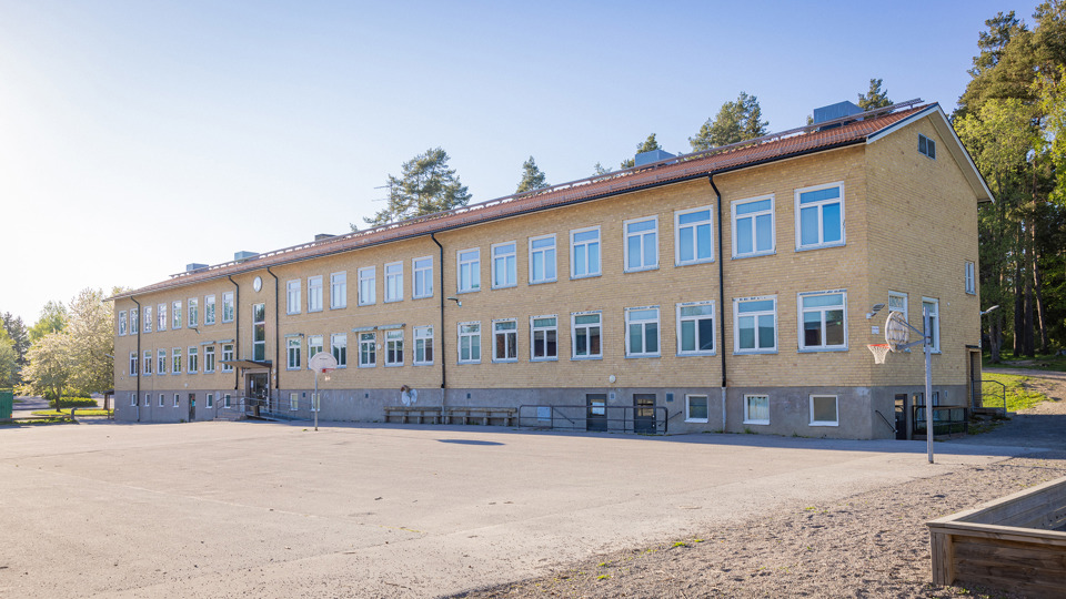 Gul tegelskola med grusplan och basketkorgar i förgrunden.