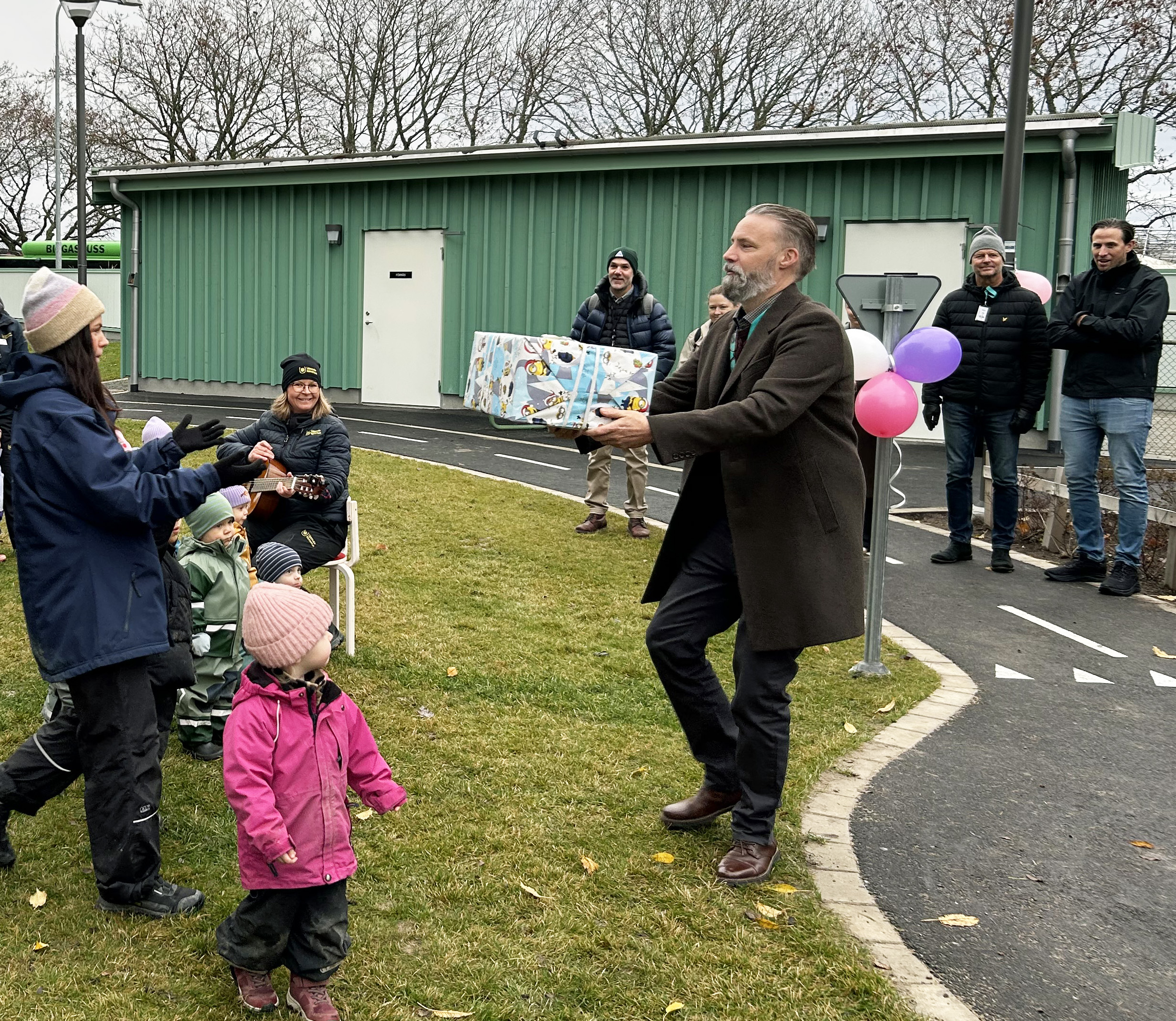 En man ger ett paket till en kvinna utomhus, omgiven av barn och vuxna. Ballonger hänger på en stolpe.