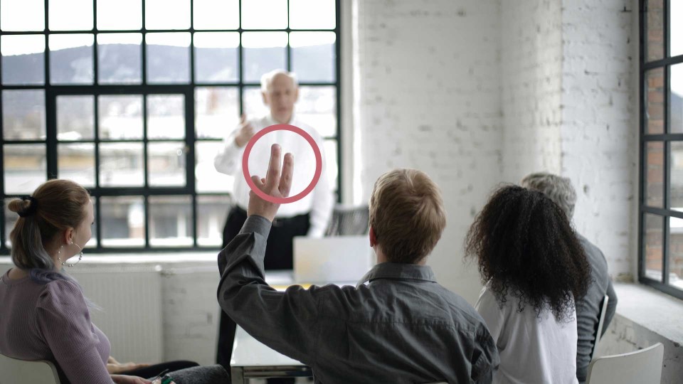 Fem personer runt ett bord, en av personerna räcker upp handen. 