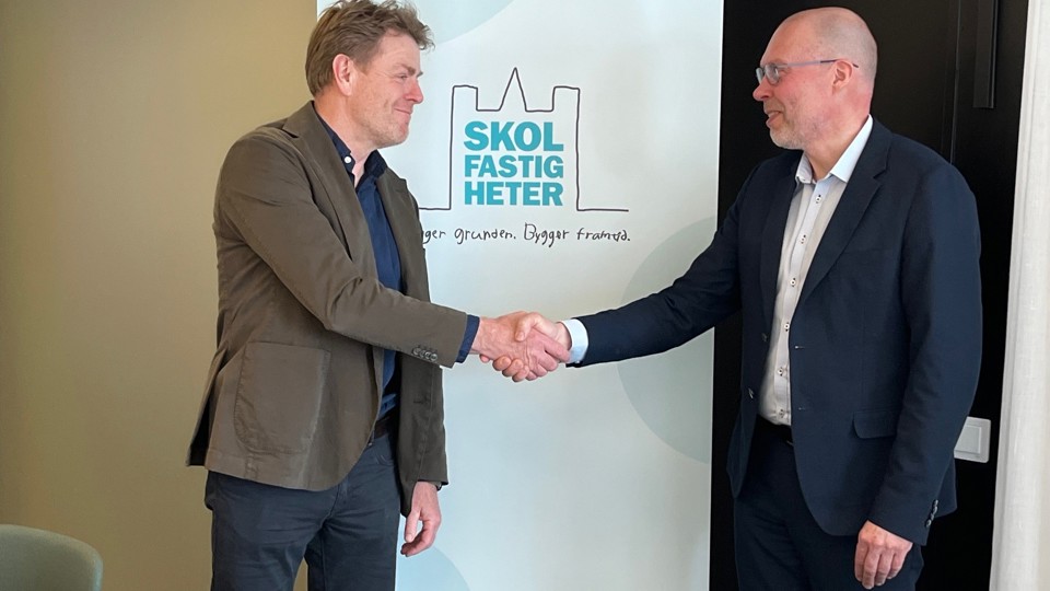 Två personer skakar hand på ett kontor framför en rollup med Skolfastigheters logotyp