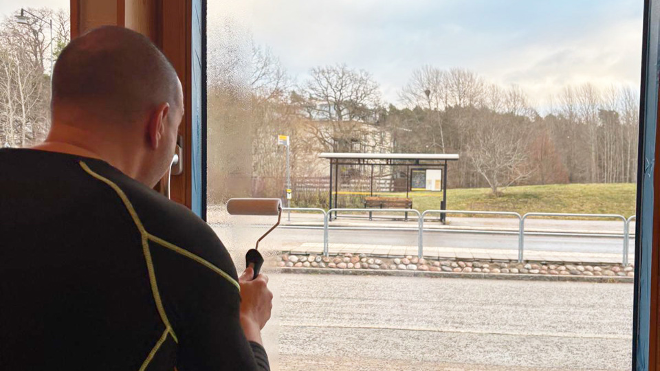 En man använder en roller för att måla en osynlig lösning på ett klassrumsfönster.