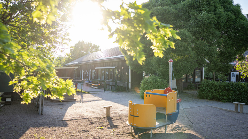 Brun träförskola med förskolegård och klätterställning i förgrunden.