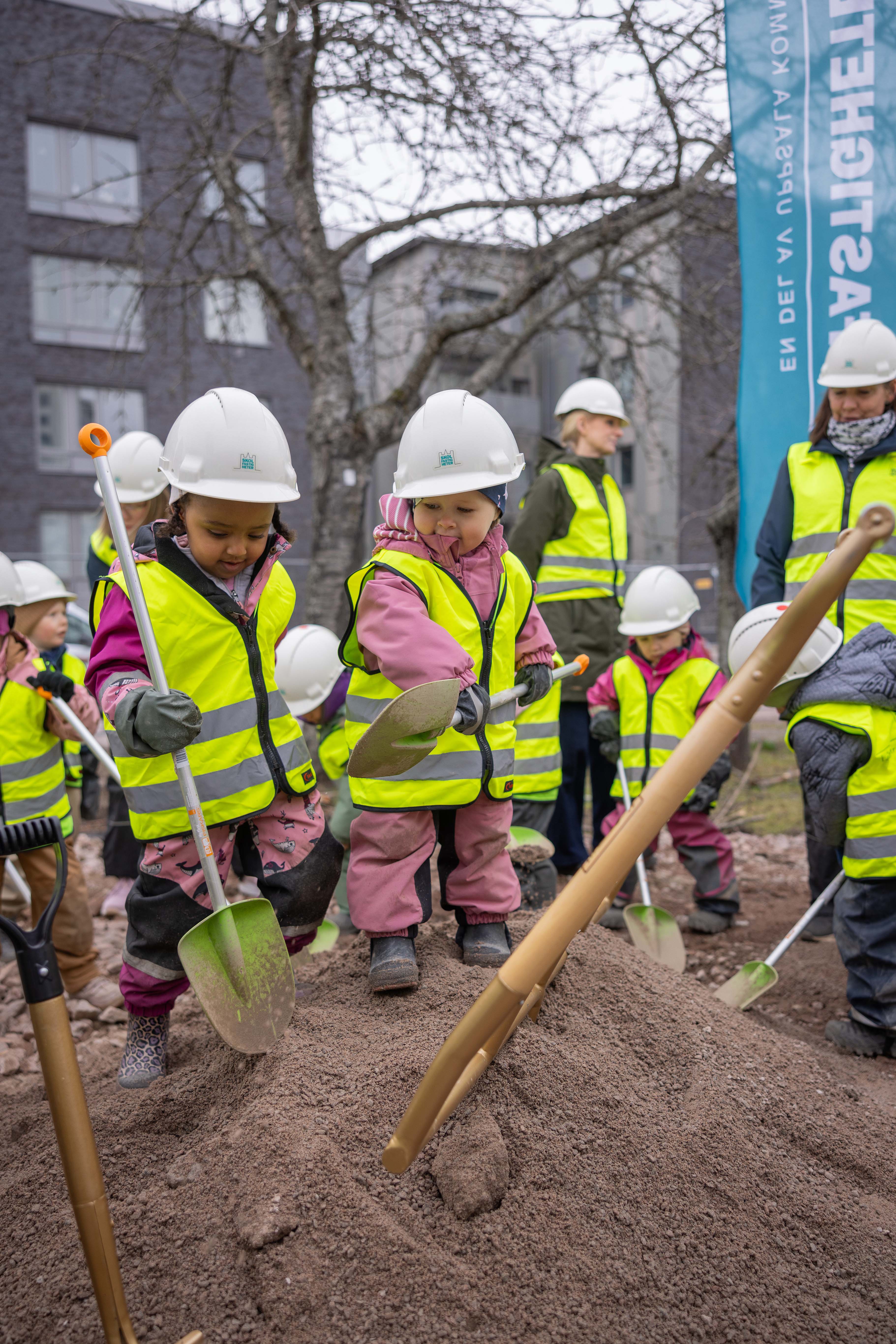 Barn i gula västar och bygghjälmar gräver i en sandhög.