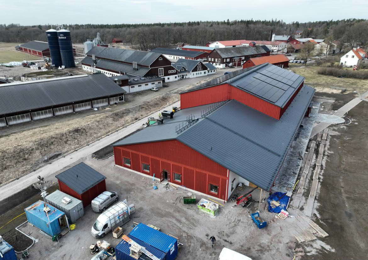 Röd byggnad belägen i lantlig lantbruksmiljö med ladugårdar och hus i bakgrunden.