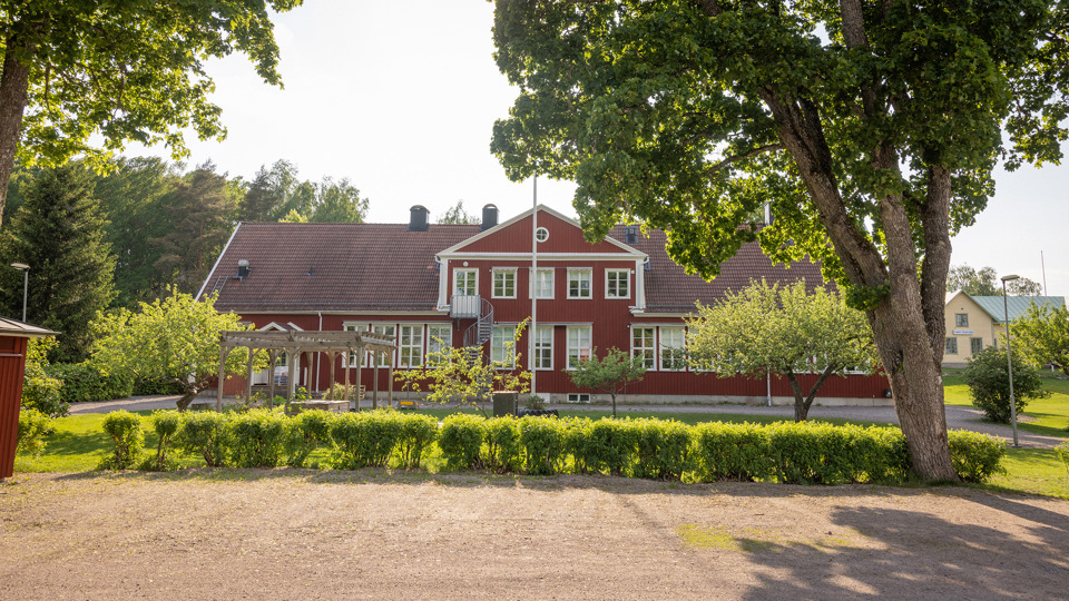 Röd skola i trä med gröna buskar och träd i förgrunden.